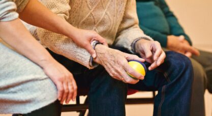 An elderly person receives support from a caregiver, holding hands indoors, showcasing compassion.